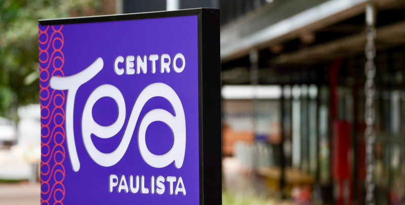 fotografia colorida de placa roxa com o logo do Centro TEA Paulista, onde a palavra TEA é formada por um símbolo do infinito desenhado pelas letras E e A. Ao fundo, desfocado, prédio do Centro TEA Paulista