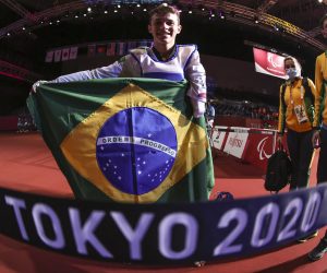 Nathan está em pé após prova, segurando uma bandeira do brasil aberta, ele veste roupa branca e está sorrindo, logo abaixo da imagem o destaque para o campeonato "TOKYO 2020"