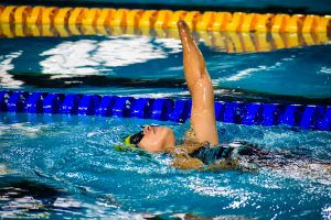 atleta em piscina durante prova, ela está nadando costas com um dos braços levantados