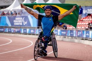 Cristian está em pista de atletismo em sua cadeira de rodas adaptada para corrida, ele está com os dois braços abertos segurando a bandeira do Brasil.