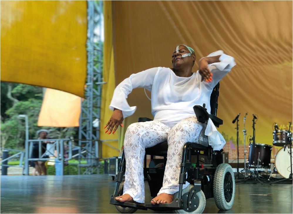Mulher negra de roupas brancas sentada em cadeira de rodas preta dança durante programação da Virada Inclusiva