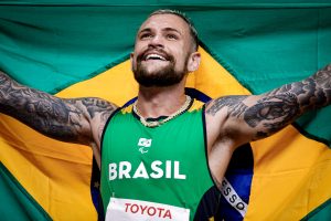 Fotografia colorida de Vinícius Rodrigues, homem de pele branca e cabelos curtos loiros, eles está com os braços levantados segurando uma bandeira do Brasil atrás do corpo, ele está olhando para cima e sorrindo.