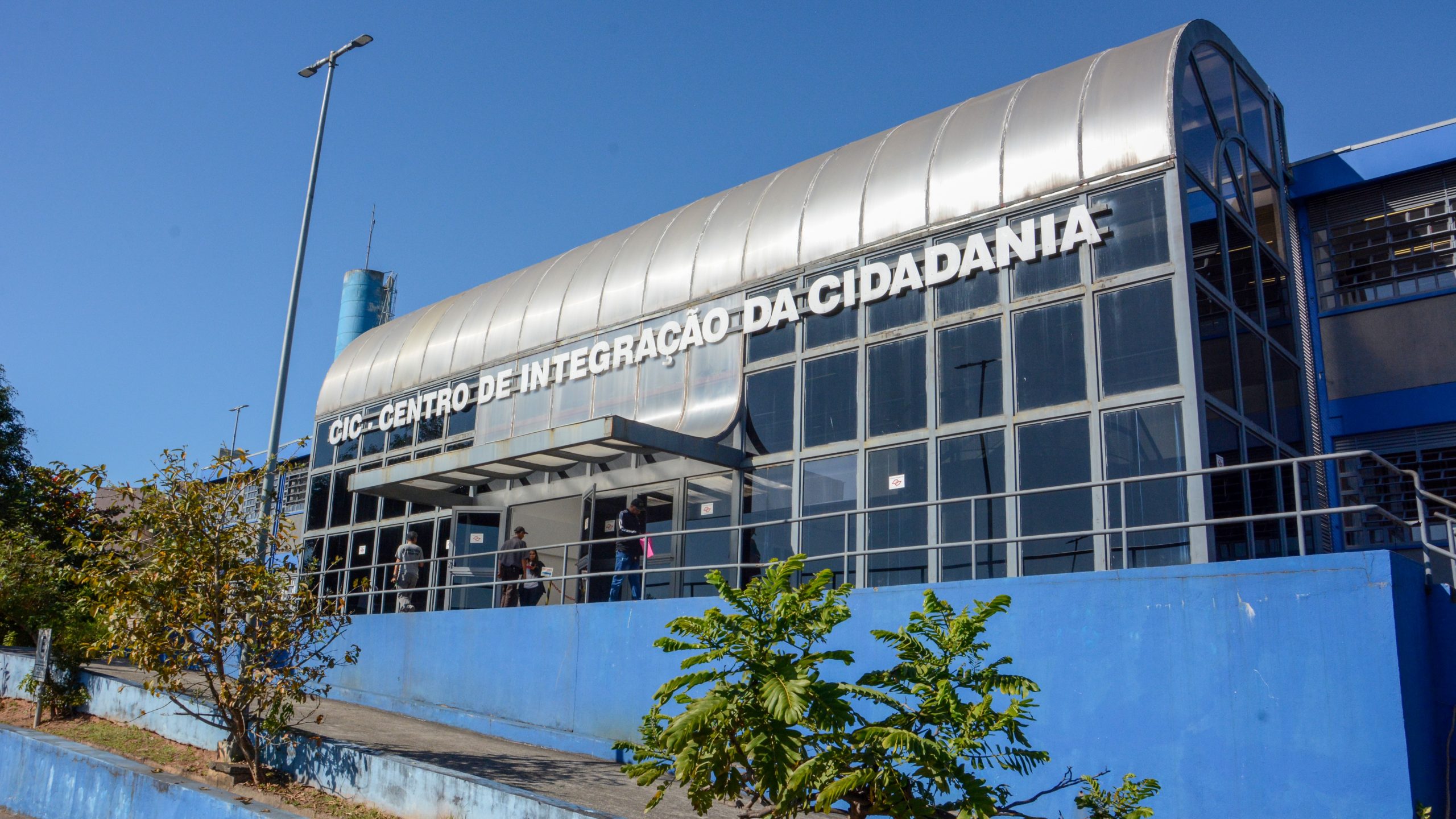 A foto mostra o Centro de Integração da Cidadania de Ferraz de Vasconcelos, em São Paulo. Está de dia, o céu está nublado com nuvens cinzas, a parede é azul e tem algumas árvores. O prédio tem vidros pretos.